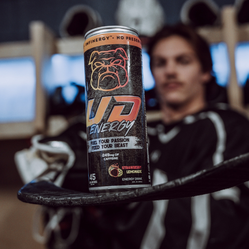 A can of UD Energy performance drink in Strawberry Lemonade flavor is in sharp focus, resting on a hockey stick&